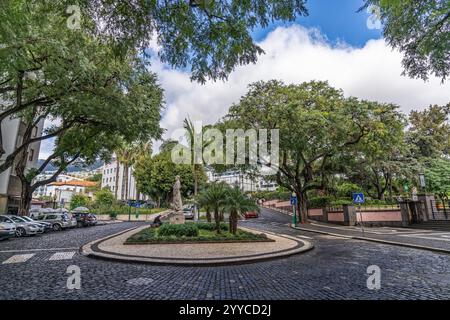Städtische Straßenlandschaft mit einem kreisrunden Kreisverkehr, umgeben von Bäumen, einer Steinstatue und einem lebhaften Stadtambiente unter teilweise bewölktem Himmel Stockfoto