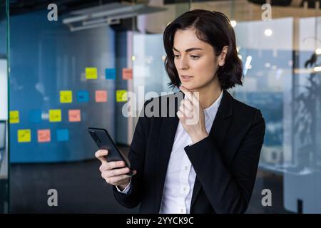 Selbstbewusste Geschäftsfrau im modernen Büro mit Smartphone für strategische Planung. Sie wirkt nachdenklich und konzentriert inmitten farbenfroher Haftnotizen, die auf Projektmanagement und Zusammenarbeit hinweisen. Stockfoto
