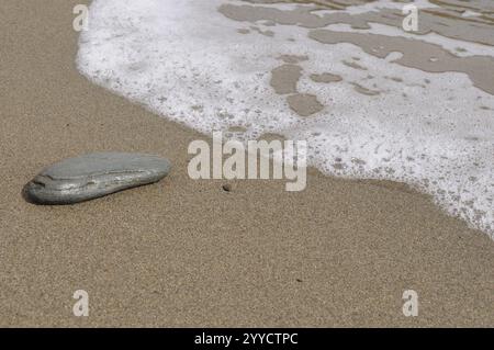 Ein ruhiger Strand mit einer hellen Sandoberfläche und sanften Wellen, die das Ufer treffen. Ein einziger Stein liegt im Sand, Spanien, Europa Stockfoto