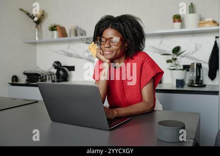 Attraktive afroamerikanische Hausfrau, die sich an die Bar lehnt, während sie online einkaufen und ihre Kreditkarte fest hält Stockfoto