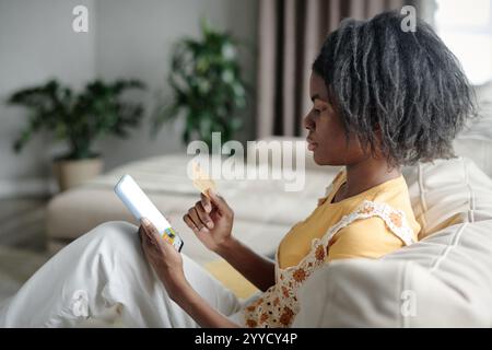 Seitenansicht einer seriösen afroamerikanischen Frau, die auf einer bequemen Couch sitzt und sich auf Online-Shopping mit goldener Kreditkarte in der Hand konzentriert Stockfoto