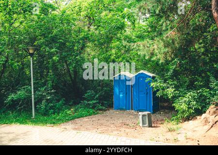 Zwei blaue, tragbare Toiletten in der Nähe von üppigem Grün, mit einem Abfalleimer davor, die wichtige Einrichtungen für Parkbesucher bieten Stockfoto