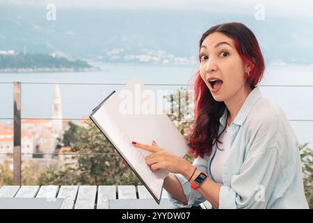Eine junge Frau mit einem Lächeln, die gerne Essen auswählt, während sie die Speisekarte in einem Restaurant am Meer mit einer wunderschönen Aussicht liest Stockfoto