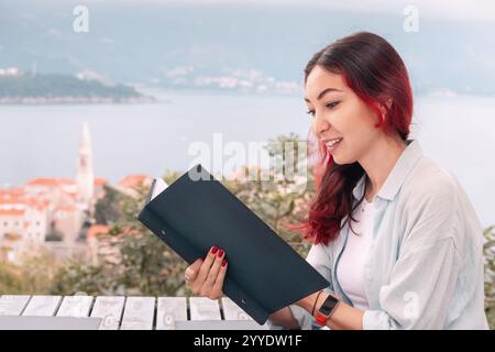 Eine junge Frau mit einem Lächeln, die gerne Essen auswählt, während sie die Speisekarte in einem Restaurant am Meer mit einer wunderschönen Aussicht liest Stockfoto