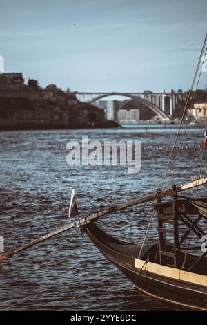 Eine vertikale Aufnahme eines traditionellen hölzernen Bootes, das am Fluss Douro mit verschwommenem Stadtbild von Porto und einer berühmten Bogenbrücke im Hintergrund unter s angedockt ist Stockfoto