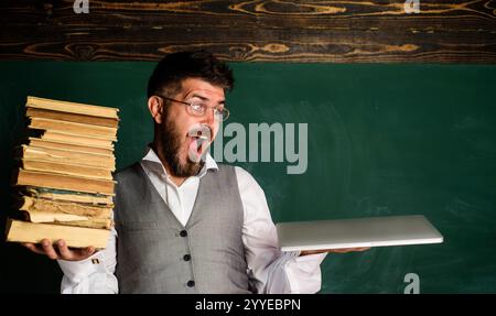 Überraschter männlicher Lehrer oder Schüler mit traditionellen Lehrbüchern und Laptop im Klassenzimmer. Elektronische Bibliothek. Moderne Bildung. Papierbuch gegen E-Book. Stockfoto