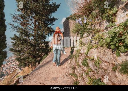 Frau, die einen Hut und einen Rucksack trägt, geht auf einem unbefestigten Pfad entlang einer Steinmauer mit Blick auf die Bucht von Kotor in Montenegro Stockfoto