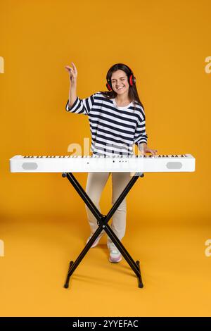 Lächelnde Frau in Kopfhörern, die Synthesizer auf orangem Hintergrund spielt Stockfoto