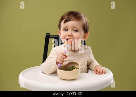 Niedliches kleines Baby, das gesundes Essen aus der Schüssel im Hochstuhl auf olivem Hintergrund isst Stockfoto