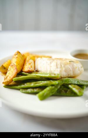 Eine Mahlzeit mit zartem gegrilltem Fisch, knusprigen gerösteten Kartoffeln und leuchtenden grünen Bohnen auf einem weißen Teller. Eine kleine Schüssel mit Soße begleitet die Stockfoto