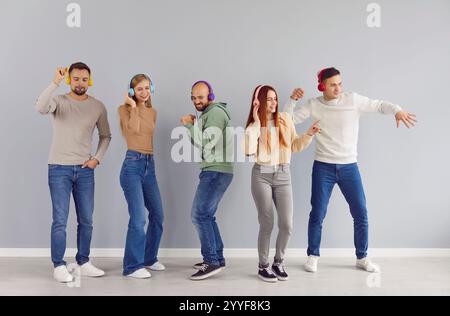 Fröhliche junge Leute in Kopfhörern, die zusammen tanzen und Musik genießen, vor grauem Hintergrund. Stockfoto