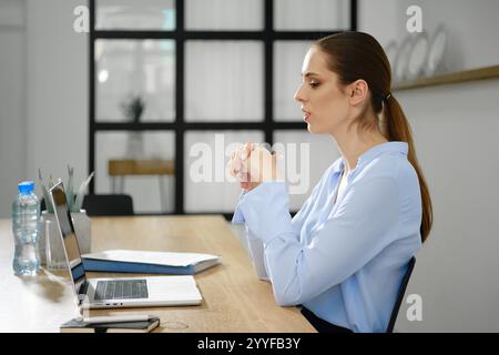 Eine Frau in einer hellblauen Bluse sitzt an einem modernen Schreibtisch, der an einem virtuellen Meeting teilnimmt, und konzentriert sich, während ihr Laptop vor ihr offen ist. Stockfoto