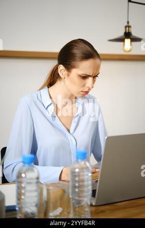 Eine junge Frau in einer blauen Bluse konzentriert sich auf ihren Laptop, während sie an einem hölzernen Schreibtisch sitzt. Zwei Wasserflaschen stehen neben ihr und Minima Stockfoto