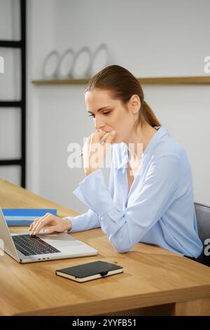 Eine junge Frau in einem hellblauen Hemd konzentriert sich auf ihren Laptop, während sie an einem hölzernen Schreibtisch sitzt. Sie scheint tief in Gedanken zu sein und ruht ihr Kinn auf Stockfoto