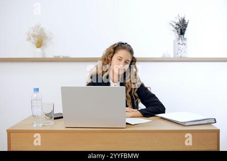 Eine junge Frau sitzt an einem modernen Holztisch und arbeitet an ihrem Laptop. Papier und eine Wasserflasche werden in der Nähe platziert, um einen aufgeräumten Arbeitsplatz zu schaffen Stockfoto