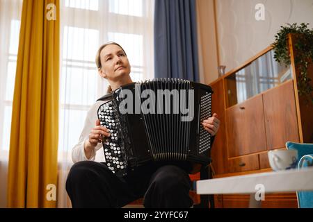 Eine Frau mittleren Alters sitzt bequem in einem gut beleuchteten Wohnzimmer und spielt leidenschaftlich Akkordeon. Sonnenlicht strömt durch das Fenster, verbessert Stockfoto