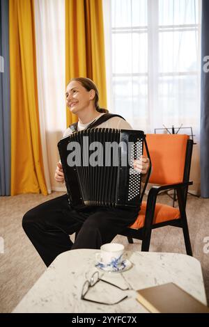 Eine Frau sitzt bequem in einem stilvollen Stuhl und lächelt, während sie die Akkordeon spielt und den gemütlichen Raum mit Musik erfüllt. Helle Vorhänge sorgen für eine gute Stimmung Stockfoto