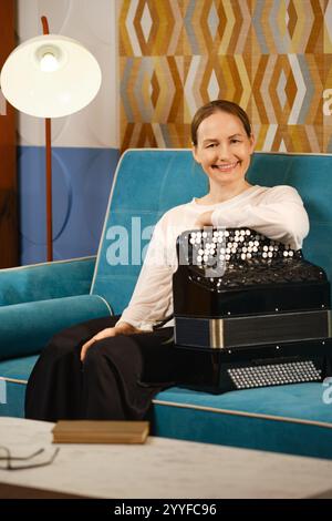 Eine Frau mittleren Alters lächelt, während sie ihr Akkordeon auf einer blauen Couch in einem gemütlichen Wohnzimmer spielt, umgeben von modernem Dekor. Stockfoto