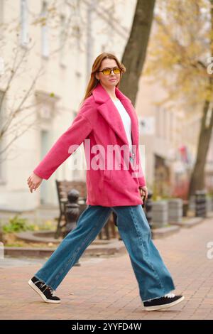 Eine Person geht selbstbewusst auf einer von Bäumen gesäumten Straße entlang, trägt einen rosa Mantel, eine Sonnenbrille und Jeans mit breiten Beinen. Das warme Herbstwetter verstärkt die che Stockfoto