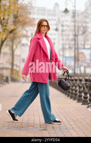 Eine stilvolle Frau schlendert in einem rosa Mantel und einer Sonnenbrille durch die Straßen der Stadt. Die herbstliche Landschaft zeigt Bäume mit gelben Blättern und einem Kopfsteinpflaster Stockfoto