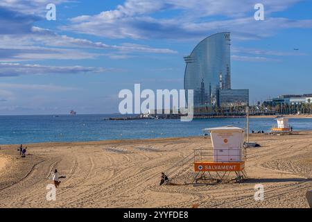 Sonniger Strand in Barcelona mit dem berühmten W Hotel, einem Rettungsschirm-Turm und einem ruhigen Mittelmeer unter einem leuchtend blauen Himmel Stockfoto