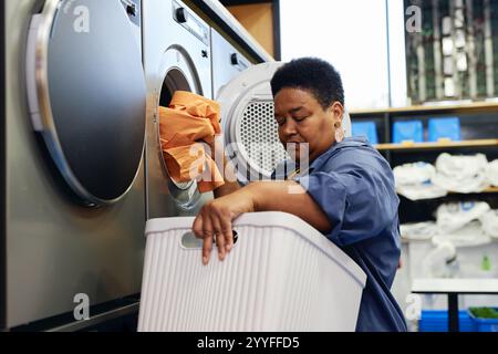 Mittelgroßer Schuss einer älteren schwarzen Frau, die Kleidung aus dem Korb in die Waschmaschine mit Münzwaschsalon in der Selbstbedienungswäsche lädt Stockfoto