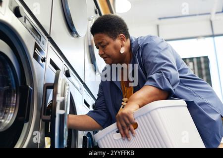 Mittelgroße Aufnahme einer älteren afroamerikanischen Frau mit Wäschekorb in den Händen, die Kleidung in die Waschmaschine in einem münzbetriebenen Waschsalon lädt, Hausarbeit-Konzept Stockfoto