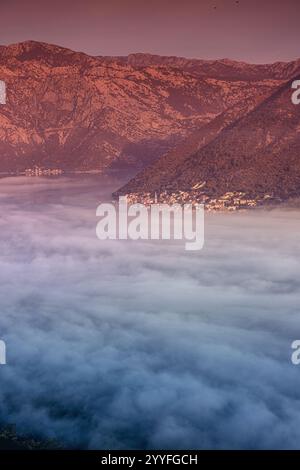 Malerischer Blick auf das Dorf Perast, das bei Sonnenaufgang teilweise von Nebel bedeckt ist, mit seinen charakteristischen roten Dächern und Kirchtürmen in der Bucht von Kot Stockfoto