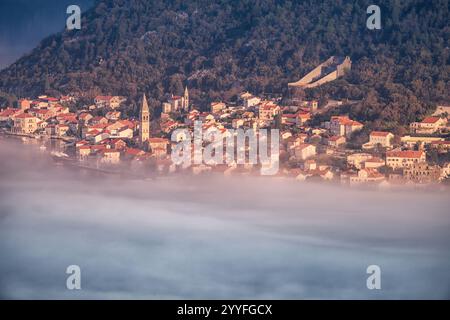 Malerischer Blick auf das Dorf Perast, das bei Sonnenaufgang teilweise von Nebel bedeckt ist, mit seinen charakteristischen roten Dächern und Kirchtürmen in der Bucht von Kot Stockfoto