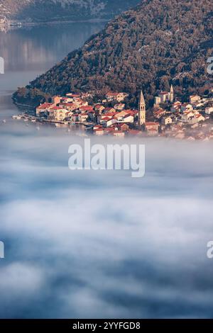Malerischer Blick auf das Dorf Perast, das bei Sonnenaufgang teilweise von Nebel bedeckt ist, mit seinen charakteristischen roten Dächern und Kirchtürmen in der Bucht von Kot Stockfoto