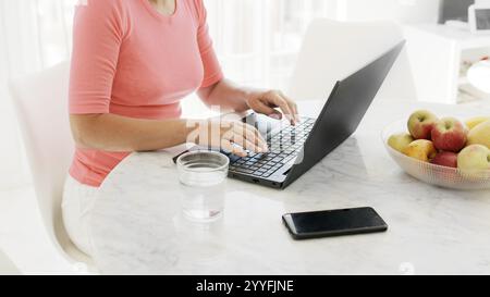 Frau mittleren Alters sitzt am Tisch und arbeitet zu Hause an einem Laptop. Schließen Sie Hände und Computer. Stockfoto