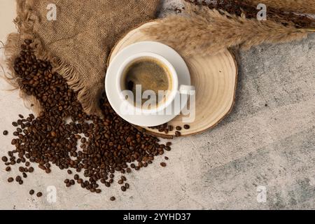 Eine Tasse Americano-Kaffee mit einer reichhaltigen Crema-Schicht - die Tasse und Untertasse wird auf eine Scheibe aus natürlichem Baumschnitt von oben, verschütteten Bohnen und Flora gelegt Stockfoto