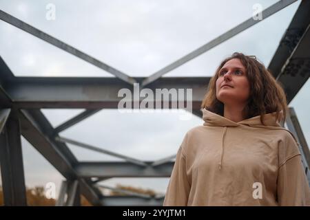 Eine erwachsene Frau steht selbstbewusst auf der Betonkonstruktion einer Industriebrücke, umgeben von schweren Balken. Eine Person steht draußen auf einem robusten i Stockfoto