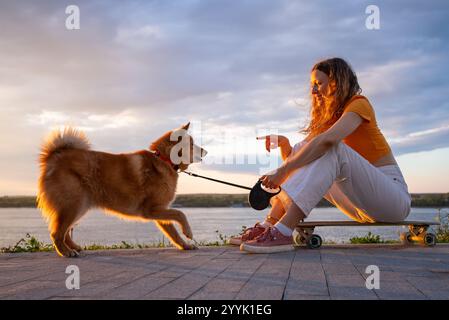 Junge Frau trainiert ihren karelischen Bärenhund, während sie bei Sonnenuntergang auf einem Skateboard am Fluss sitzt und einen friedlichen Moment der Verbindung und des Trainings mit ihrem Haustier in der Natur genießt Stockfoto