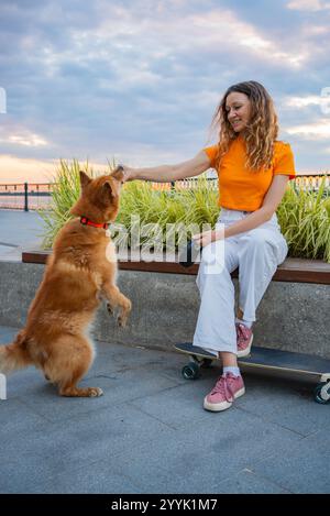 Eine junge Frau mit einem orangen T-Shirt und einer weißen Hose sitzt auf einem Skateboard und trainiert ihren karelischen Bärenhund draußen in einem Park nahe einem Fluss bei Sonnenuntergang Stockfoto