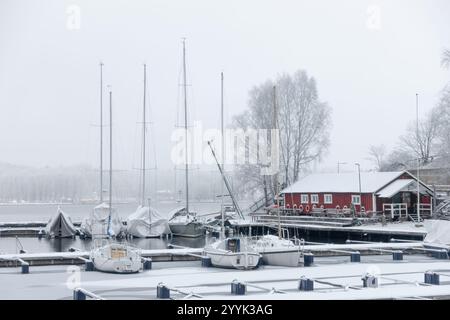 Weiße Winterszene mit kleinen Booten in einem schneebedeckten Yachthafen mit rotem Bootshaus am Ufer an einem bewölkten nebeligen Tag Stockfoto