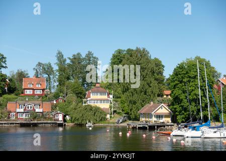 Typisch schwedische Holzhäuser mit weißen Zierleisten am Strand in idyllischer Sommerlandschaft am Ufer mit Segelbooten, die im Sommer an der Anlegestelle anlegen Stockfoto