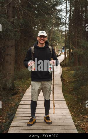 Ein Mann, der gemütlich einen rustikalen Holzsteg hinuntergeht, während er eine erfrischende Dose CocaCola in der Hand hält und seinen Spaziergang im Freien ganz klar genießt Stockfoto