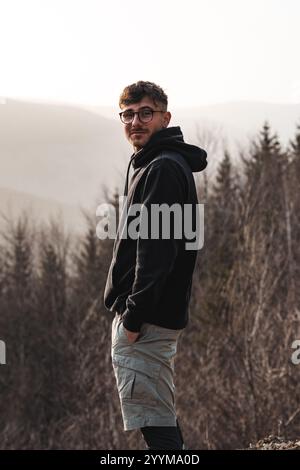 Ein Mann mit Brille und schwarzem Hoodie steht selbstbewusst vor einem üppigen Wald, der reich an Grün und Leben ist Stockfoto