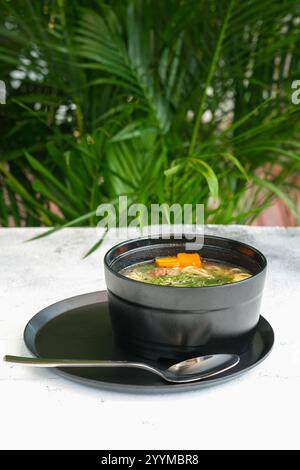 Eine wohltuende Schüssel mit klarer Suppe mit Karotten, Gemüse und Kräutern, elegant serviert in einer schwarzen Keramikschale auf einem passenden Teller Stockfoto