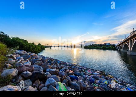 Eine Brücke überspannt einen Fluss mit einem wunderschönen Sonnenuntergang im Hintergrund. Das Wasser ist ruhig und der Himmel ist klar Stockfoto