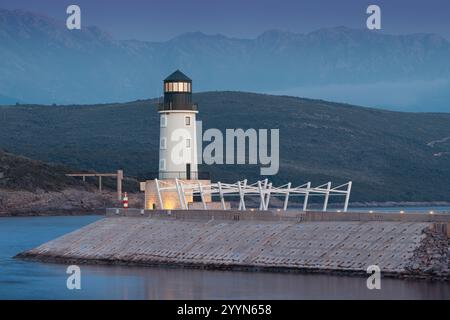 Ruhige Küstenlandschaft mit einem weißen Leuchtturm an einem Pier, vor einem Hintergrund der Berge bei einem lebhaften Sonnenuntergang Stockfoto