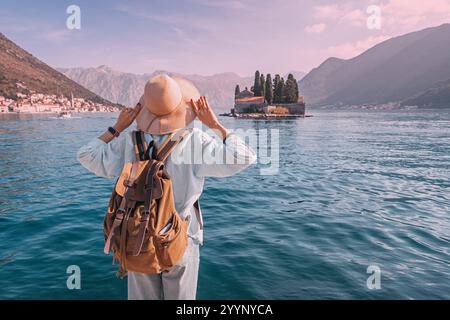 Weibliche Touristen mit Rucksack und Hut, die Arme heben, genießen die malerische Aussicht auf die Insel mit Kloster in der Bucht von Kotor, Perast, Montenegro Stockfoto