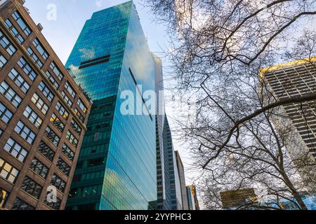 Moderner Glashochhaus, der Himmel und Wolken reflektiert, umgeben von blattlosen Bäumen und benachbarten historischen Gebäuden in New York City. Stockfoto