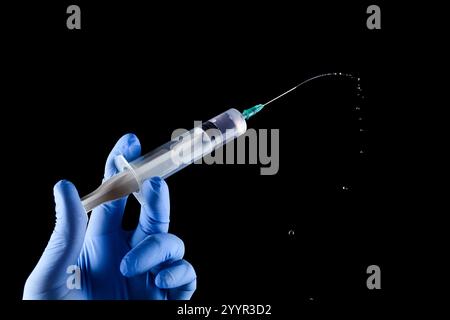 Hand in medizinische Schutzhandschuhe, die eine Spritze mit Medikamenten und Injektionstropfen halten, isoliert auf schwarzem Hintergrund in einem Studio. Kopierbereich Stockfoto