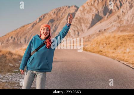 Junge Reiseteilnehmerin, die auf einer Bergstraße trampelt und vorbeifahrenden Autos zuwinkt Stockfoto