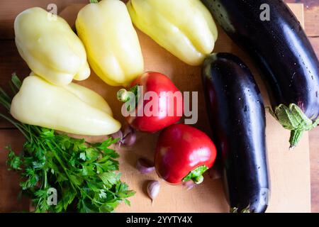 Bunte Auswahl an frischem Gemüse mit gelben und roten Paprika, Auberginen, Knoblauch und Koriander, alles ordentlich auf einer hölzernen Schneideboa angeordnet Stockfoto