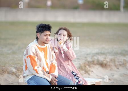 Ein Paar im Alter von 20 Jahren isst im Herbst in Tokoname, Präfektur Aichi, gerne Crepes auf einer kalten Strandbank. Stockfoto