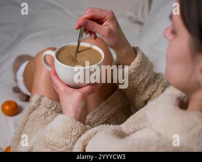 Frau im Bademantel sitzt auf weißer Decke und rührt eine Tasse Kaffee mit Löffel um. Sie trägt warme Socken Stockfoto