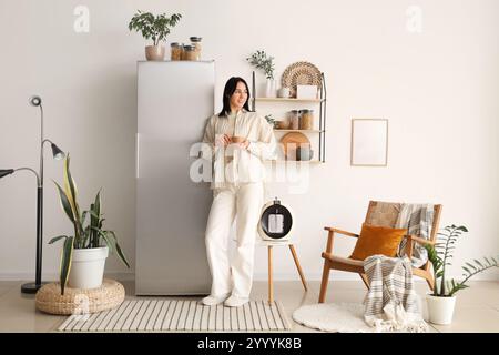 Schöne junge Frau mit einer Tasse Kaffee, die in der Nähe eines modernen Kühlschranks in einer stilvollen Küche steht Stockfoto